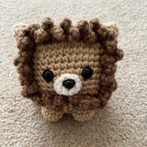 Puede incluir: Un león de crochet con pelo marrón, ojos negros y nariz blanca. El león es un pequeño animal de peluche.