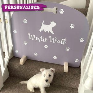 May include: A personalised, lilac-coloured pet gate with a bone-shaped handle and paw print accents. The gate features a white silhouette of a West Highland White Terrier and the text "Westie Wall". A small white Westie puppy sits in front of the gate.