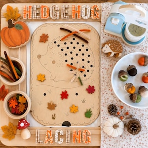 Puede incluir: Un tablero de madera para enhebrar con forma de erizo y agujeros para limpiapipas, con el texto HEDGE HOG LACING. Incluye una calabaza de madera, una seta, una figura de erizo, hojas de fieltro, bellotas, una cafetera de juguete y piñas.