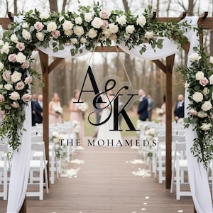 May include: Wedding ceremony setup with a wooden arch decorated with white and blush roses, greenery, and white fabric. A clear sign with the initials "A&K" and the name "THE MOHAMEDS" hangs from the arch. White chairs line the aisle.