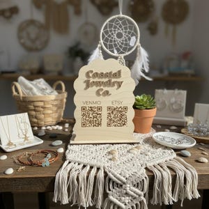 May include: A wooden sign for "Coastal Jewelry Co." with QR codes for Venmo and Etsy. The sign is on a table with jewelry, a macrame runner, and a small potted succulent. A dreamcatcher hangs in the background.