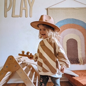 May include: A young child wearing a brown hat and striped sweater plays with a wooden toy. The child is standing on a wooden floor in front of a wooden climbing structure. The word "PLAY" is written on the wall behind the child. A rainbow-colored wall hanging is visible in the background.