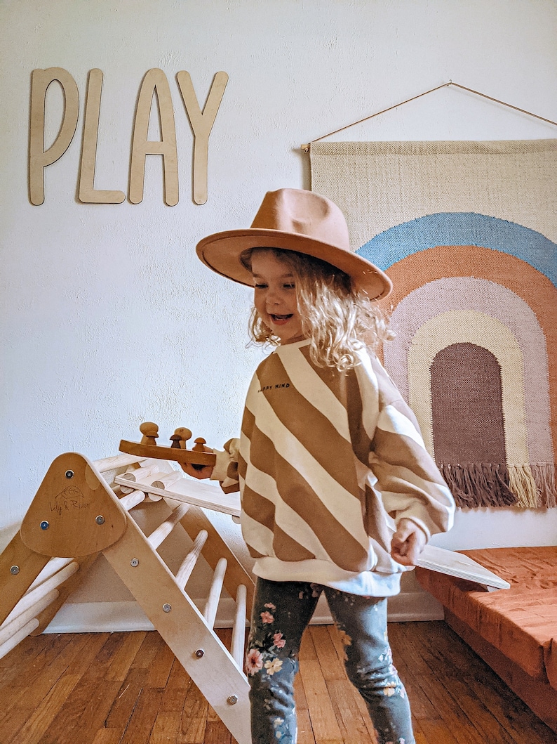 May include: A young child wearing a brown hat and striped sweater plays with a wooden toy. The child is standing on a wooden floor in front of a wooden climbing structure. The word "PLAY" is written on the wall behind the child. A rainbow-colored wall hanging is visible in the background.