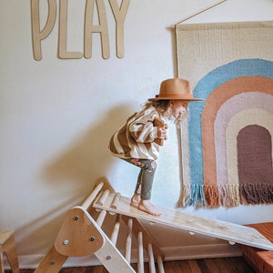 May include: A child wearing a brown and white striped sweater, brown pants, and a brown hat is playing on a wooden climbing triangle with a slide. The word "PLAY" is written on the wall behind the child. A rainbow-colored wall hanging is on the wall to the right of the child.