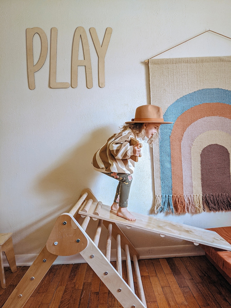 May include: A child wearing a brown and white striped sweater, brown pants, and a brown hat is playing on a wooden climbing triangle with a slide. The word "PLAY" is written on the wall behind the child. A rainbow-colored wall hanging is on the wall to the right of the child.