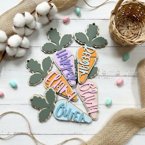 May include: Wooden carrot-shaped Easter ornaments with names in various colors. Each carrot has a green leaf design and a name in a different color, such as purple, orange, and blue. The background includes cotton stems, a basket, and Easter egg candies.