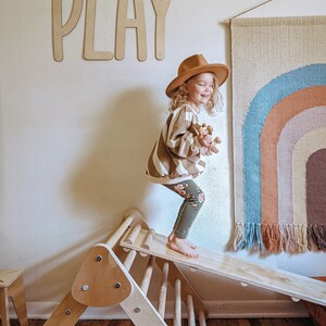 May include: A young child wearing a brown hat and a striped sweater is playing on a wooden climbing triangle and slide. The word "PLAY" is written on the wall behind the child. A rainbow wall tapestry hangs on the wall to the right of the child.