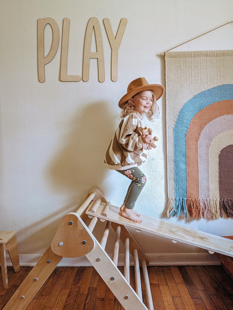 May include: A young child wearing a brown hat and a striped sweater is playing on a wooden climbing triangle and slide. The word "PLAY" is written on the wall behind the child. A rainbow wall tapestry hangs on the wall to the right of the child.