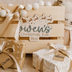 May include: A wooden Christmas Eve box with the name "Owen's" in blue script, decorated with a Santa and reindeer design. The box is surrounded by wrapped gifts with name tags and a garland of white pom-poms.