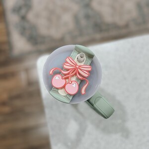 May include: A green and white tumbler lid with a pink cherry and bow design. The lid has a straw hole and a silicone seal.