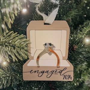 May include: A wooden ornament shaped like a ring box with a diamond ring inside. The box is painted a light brown color and has the word "engaged" written on it in black cursive. The year "2024" is written below the word "engaged".