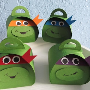 May include: Four green Teenage Mutant Ninja Turtles themed party favor boxes with different colored masks. Each box has a handle, googly eyes, and a smiling mouth.