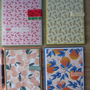 May include: Four notebooks with fruit-themed designs. One has a watermelon pattern with a watermelon ribbon closure and the text "LIFE IS SWEET". Another features pineapples, and the others have peach and orange patterns. A pen rests on the peach notebook.
