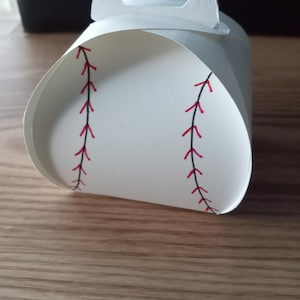 May include: A white baseball-themed gift box with a handle. The box is cylindrical with red stitching details, mimicking a baseball. The handle is a curved, rectangular shape. The box is made of paper.