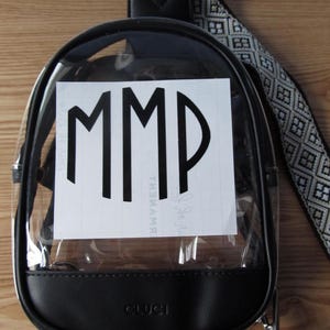 May include: Clear, rounded backpack with black trim and a patterned strap. The backpack features the letters "MMP" in bold, black font on a white background. The bottom of the backpack has the word "CLUCI" printed on it.