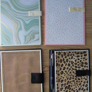 May include: Four notebooks with different cover designs. One has a teal and gold marble pattern, another has a light blue background with small colorful dots, a third has a brown leather-like texture, and the last has a leopard print. Each has a closure.
