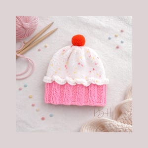 May include: A handmade knitted hat in the shape of a cupcake. The hat features a pink ribbed base, a white top with colorful speckles, and a white ruffled edge. An orange pom-pom tops the hat. Knitting needles and yarn are in the background.