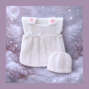 May include: A white knitted baby outfit set, comprising a dress with pink buttons and a matching hat. The dress has a small white bow and decorative stitching. The set is designed for newborns and infants.
