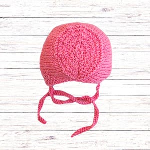 Puede incluir: Un gorro tejido de color rosa brillante que presenta un patr&oacute;n circular texturizado en la coronilla y lados de punto liso. La prenda incluye dos lazos largos de hilo trenzado unidos en la base para asegurar bajo la barbilla.
