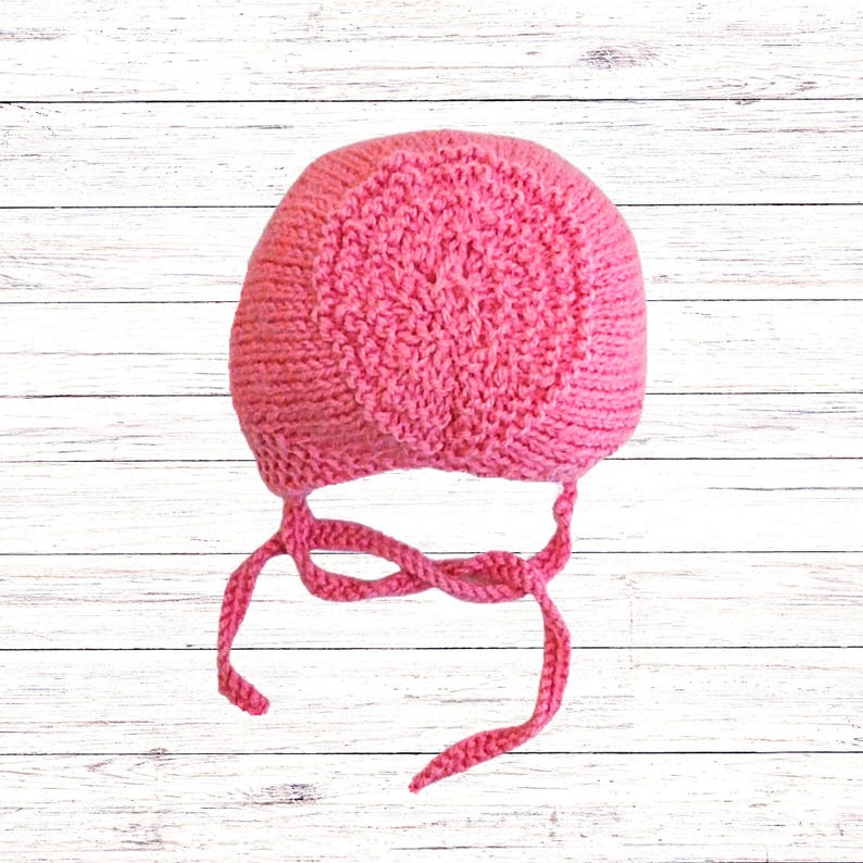 Puede incluir: Un gorro tejido de color rosa brillante que presenta un patr&oacute;n circular texturizado en la coronilla y lados de punto liso. La prenda incluye dos lazos largos de hilo trenzado unidos en la base para asegurar bajo la barbilla.