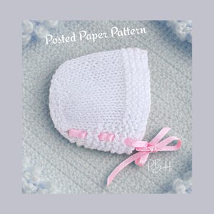 May include: A white knitted baby bonnet with pink ribbon detailing and a bow. The bonnet is presented on a light blue knitted blanket. The text "Posted Paper Pattern" is visible at the top.