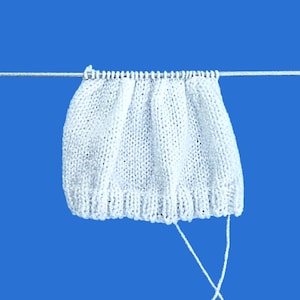 May include: A white knitted fabric piece, possibly part of a garment, hangs against a blue background. The knitted item has a textured appearance with visible stitches and a scalloped edge. The yarn is white, and the piece is suspended by a thin, white thread.