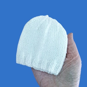 May include: A white knitted beanie hat. The hat is held in a hand against a blue background. The hat is made of a soft, textured yarn and has a ribbed brim. The hat is a solid colour and appears to be handmade.