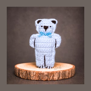Puede incluir: Un oso de peluche blanco hecho a mano con detalles marrones, incluyendo las orejas y la nariz. El oso tiene una pajarita azul claro y está sobre una pequeña rodaja de madera redonda. El fondo es de color marrón liso.