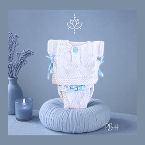 May include: A white knitted baby outfit, comprising a top and nappy cover, with light blue ribbon details. The top features a single blue button. The outfit is displayed on a light blue, round, textured cushion. A candle and vase are in the background.