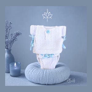 May include: A white knitted baby outfit, comprising a top and nappy cover, with light blue ribbon detailing. The top features a single blue button. The outfit is displayed on a light blue, round, textured cushion. A candle and vase are in the background.