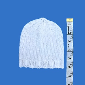 May include: A white knitted beanie hat, approximately 15 cm in height, is displayed against a blue background. The hat is made of a soft, textured yarn, with a ribbed brim. A measuring tape is placed next to the hat.