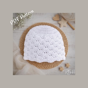 May include: A white crocheted hat with a textured pattern, on a round woven mat. The image includes the text "PDF Pattern" and wooden shapes. The hat is a handmade item.