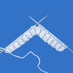May include: A close-up of white knitted fabric with two silver knitting needles inserted. The knitted piece is in a cable stitch pattern, against a blue background. The yarn is white.