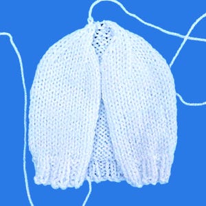 May include: A white, hand-knitted baby cardigan with a rounded neckline and long sleeves. The cardigan is made of soft yarn and has a delicate texture. White yarn strands extend from the top of the cardigan. The background is a solid blue.
