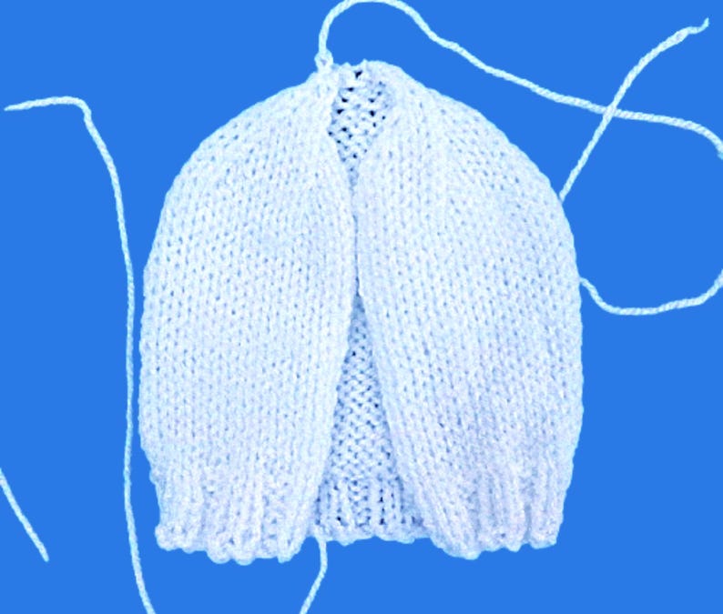 May include: A white, hand-knitted baby cardigan with a rounded neckline and long sleeves. The cardigan is made of soft yarn and has a delicate texture. White yarn strands extend from the top of the cardigan. The background is a solid blue.