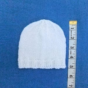 May include: A small, white knitted hat, possibly for a baby, is placed on a blue surface. A yellow and white measuring tape is next to the hat. The hat is approximately 13 cm in height.