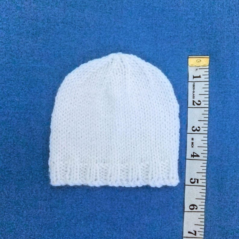 May include: A small, white knitted hat, possibly for a baby, is placed on a blue surface. A yellow and white measuring tape is next to the hat. The hat is approximately 13 cm in height.