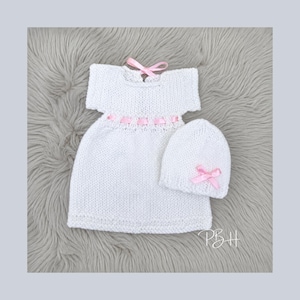 May include: A white knitted baby dress and matching hat set. The dress has short sleeves and a pink ribbon detail around the waist. Both items have pink bows. The set is displayed on a grey, textured surface.