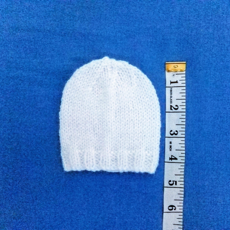 May include: A small, white knitted hat is displayed on a blue surface. A measuring tape is placed next to the hat, showing a length of approximately 15 cm. The hat has a rounded top and a ribbed brim.