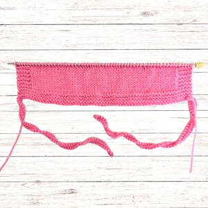 Puede incluir: Una pieza rectangular de tejido de punto rosa brillante sobre una aguja de tejer de metal larga. La pieza presenta una secci&oacute;n central en punto jersey bordeada por bordes en punto bobo, con dos lazos de punto largos y finos en las esquinas.