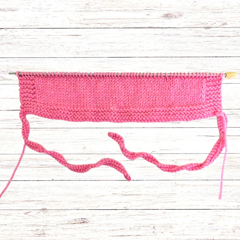 Puede incluir: Una pieza rectangular de tejido de punto rosa brillante sobre una aguja de tejer de metal larga. La pieza presenta una secci&oacute;n central en punto jersey bordeada por bordes en punto bobo, con dos lazos de punto largos y finos en las esquinas.
