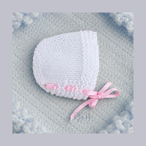May include: A white knitted baby bonnet with pink ribbon detailing and a bow. The bonnet rests on a light blue blanket with a textured pattern and a decorative border. The initials "PBH" are visible.
