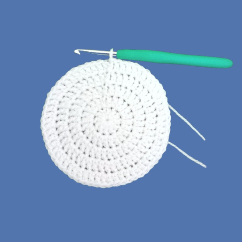 Peut inclure: Un cercle blanc crochet&eacute; avec un crochet vert sur fond bleu. Le projet de crochet est en cours, le crochet &eacute;tant ins&eacute;r&eacute; dans les mailles. Deux brins de fil blanc d&eacute;passent du cercle.