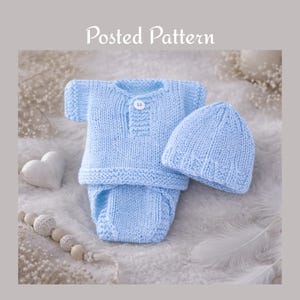 May include: A light blue knitted baby outfit set, comprising a shirt with a button fastening, a diaper cover, and a matching beanie. The words "Posted Pattern" are in the top center of the image. The set is displayed on a soft, white surface.