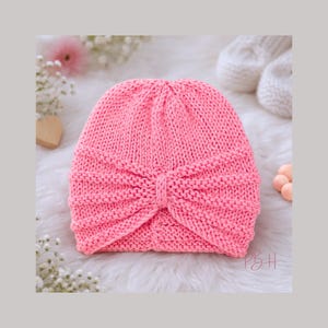 Puede incluir: Un gorro tipo turbante rosa tejido. El gorro presenta un diseño anudado en la parte delantera y está hecho de un hilo suave y texturizado. El gorro es de color rosa sólido y vibrante y probablemente está hecho a mano. El fondo es una superficie blanca y suave.