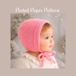 May include: A pink knitted baby bonnet with a matching tie. The bonnet is made of a soft, textured yarn and has a ribbed edge. The words "Posted Paper Pattern" are in the top center of the image.