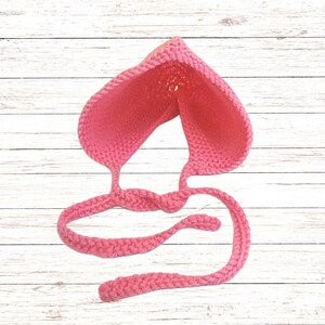 Puede incluir: Un gorro de ganchillo rosa brillante con una parte superior puntiaguda y un patr&oacute;n de punto texturizado. La prenda incluye dos lazos largos de ganchillo trenzado que salen de la base, dise&ntilde;ados para atarse bajo la barbilla de forma segura.
