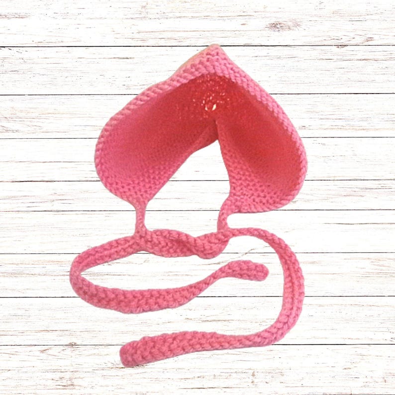 Puede incluir: Un gorro de ganchillo rosa brillante con una parte superior puntiaguda y un patr&oacute;n de punto texturizado. La prenda incluye dos lazos largos de ganchillo trenzado que salen de la base, dise&ntilde;ados para atarse bajo la barbilla de forma segura.