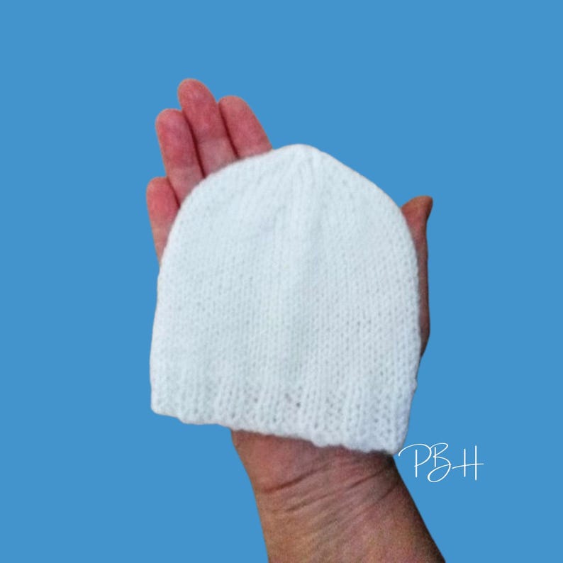 May include: A white knitted beanie hat, held in a hand, against a blue background. The hat is made of a soft, textured yarn. The initials "PBH" are written in white script in the lower right corner.