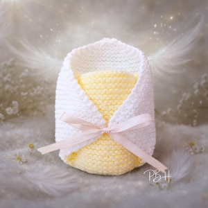 May include: A hand-knitted baby cocoon in white and yellow, tied with a light pink ribbon. The background features soft white feathers and a dreamy, blurred effect. The cocoon is a soft, cozy design.